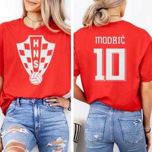 A woman wearing a red t-shirt with a white crest and the number 10 on the back, paired with distressed jeans.