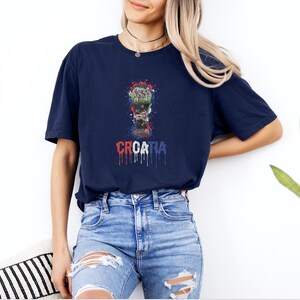 A woman wearing a navy blue t-shirt with a graphic design and the word "Cronia" printed on it, standing against a white wall.