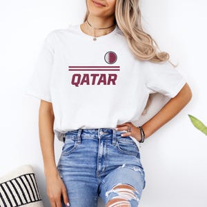 A woman wearing a white t-shirt with the word "Qatar" printed on it, paired with distressed blue jeans, stands in front of a white wall.