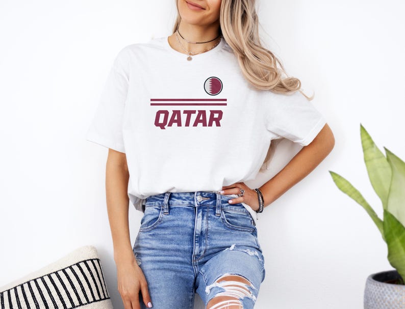 A woman wearing a white t-shirt with the word "Qatar" printed on it, paired with distressed blue jeans, stands in front of a white wall.