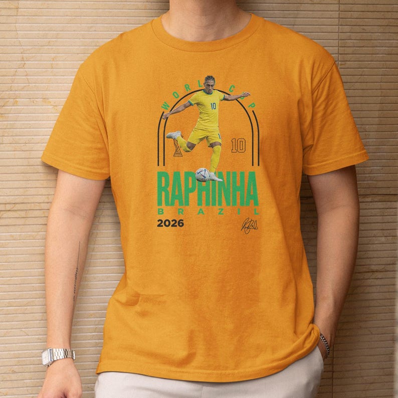 a yellow t-shirt with a graphic design of a soccer player in yellow and green colors, along with the text "RAPINHA BRASIL" and the year "2026".