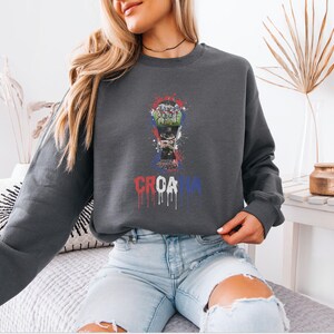 A woman wearing a gray sweatshirt with a graphic design of a soccer ball and the word "CRCATIA" printed on it, sitting on a couch.