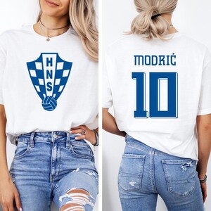 A woman wearing a white t-shirt with a blue logo and the number 10, as well as blue jeans, posing in front of a white background.
