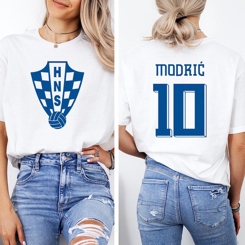 A woman wearing a white t-shirt with a blue logo and the number 10, as well as blue jeans, posing in front of a white background.