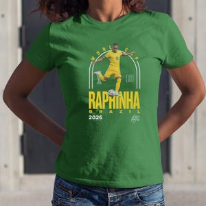 A green t-shirt with a graphic design featuring a soccer player in yellow and white, along with the text "RAPHINHA BRASIL" and the year "2026".