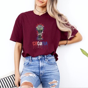 A woman wearing a burgundy t-shirt with a graphic of a trophy and the word "Croatia" printed on it, standing against a white wall.