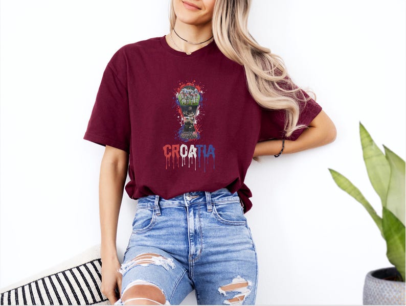 A woman wearing a burgundy t-shirt with a graphic of a trophy and the word "Croatia" printed on it, standing against a white wall.