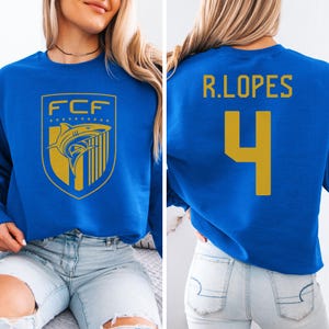 May include: Royal blue sweatshirt featuring a gold shield design with a shark graphic and the letters "FCF". The back of the sweatshirt displays the name "R.LOPES" and the number "4" in gold.
