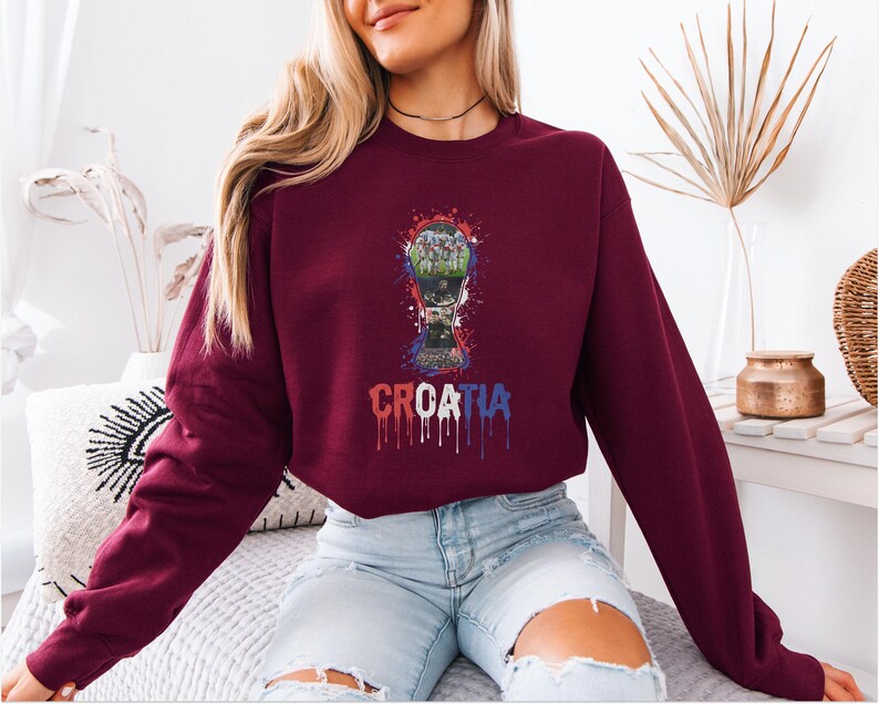 A woman wearing a burgundy sweatshirt with a graphic of a soccer ball and the word "CRETIA" printed on it, sitting on a bed.