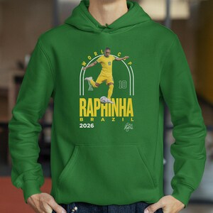 A green hoodie with a graphic design featuring a soccer player in yellow uniform and the text "RAPHINHA BRAZIL" printed on it.