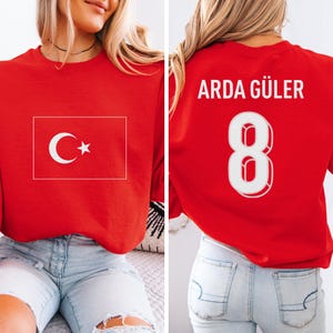 May include: Red sweatshirt featuring the Turkish flag on the front. The back of the sweatshirt displays the name "ARDA GÜLER" above the number "8" in white. The sweatshirt is a solid red color.