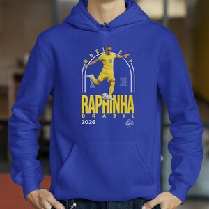 A blue hoodie with a graphic of a soccer player in yellow and white, along with text that reads "RAPHINHA BRAZIL" and "2026".
