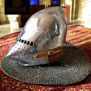 Può includere: Un elmo medievale in metallo lucido con visiera appuntita e fori di ventilazione. L'elmo ha un cinturino in pelle ed è posto su una cuffia di cotta di maglia. L'immagine include il testo "UNIQUE" e "40% OFF".