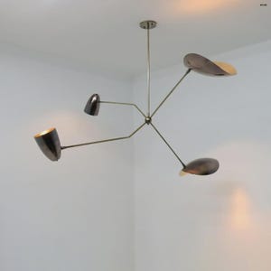 Puede incluir: Un moderno candelabro de varios brazos con acabado de latón. La lámpara presenta cuatro pantallas curvas en forma de hoja con un interior contrastante. La lámpara está suspendida de un soporte de techo.