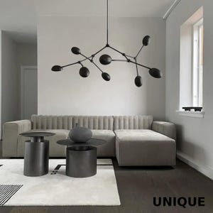 May include: A contemporary living room with a grey sofa, two dark grey side tables, and a black chandelier with oval lights. A decorative vase sits on one table. The word "UNIQUE" is visible.