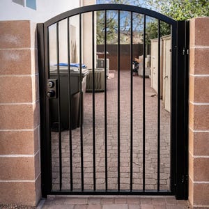 May include: A black metal gate with a curved top and vertical bars. The gate is set between two brick pillars. A black handle and lock are visible on the gate. The gate opens to a brick paved area.