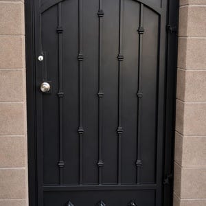 May include: A black wrought iron gate with an arched top and vertical bars. The gate features decorative diamond-shaped accents and is flanked by tan brick columns. A silver doorknob and lock are visible.
