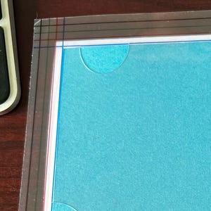 May include: A turquoise card with a clear plastic overlay featuring rounded cutouts. The overlay has the text "©Lynne LaMaster" printed in black. The card is on a white background.