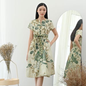 Op de afbeelding: Een beige knielange jurk met een groen en bruin bloemen- en vogelpatroon. De jurk heeft korte mouwen, een knoopsluiting aan de voorkant en een bijpassende riem. Het model draagt witte sandalen.
