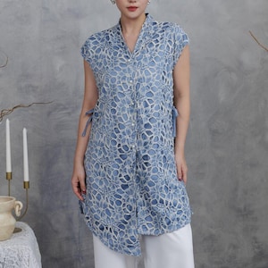May include: A light blue lace tunic with a V-neck and cap sleeves. The tunic has a button-down front and side ties. The model is wearing white pants. The background is a neutral gray.