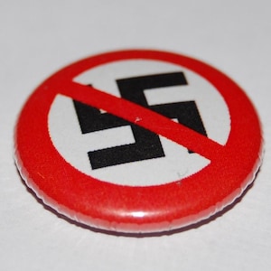 May include: A circular pinback button featuring a white background with a black swastika symbol in the centre. A thick red circular border surrounds the design, and a diagonal red line crosses through the centre of the swastika symbol.