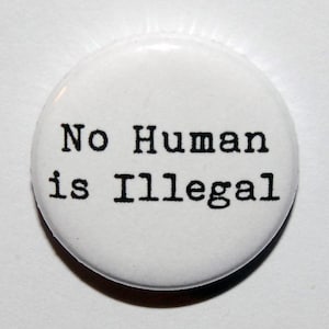 May include: A circular white pin-back button featuring the black text No Human is Illegal printed in a typewriter-style font. The button has a smooth, glossy finish and a simple, clean design with the text centred on the white background.