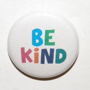 May include: A white button badge featuring the words "BE KIND" in a vibrant, multi-coloured font. The text is in uppercase letters, with each letter a different colour, including blue, green, pink, orange, and dark blue.