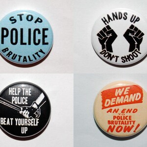 May include: Four circular badges with protest slogans. One has "STOP POLICE BRUTALITY" on a blue background. Another says "HANDS UP DON'T SHOOT" with black fists. A third says "HELP THE POLICE BEAT YOURSELF UP". The last says "WE DEMAND AN END TO POLICE BRUTALITY NOW!"
