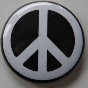 May include: A circular badge featuring a white peace symbol against a black backdrop. The peace symbol is a white line drawing within a circle. The badge has a shiny, reflective surface.
