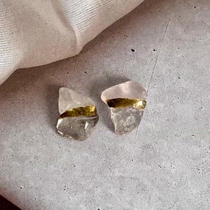 May include: A pair of earrings. Each earring is made from a clear quartz crystal with a gold-coloured band. The top of each crystal has a soft pink tint. The earrings are displayed on a grey surface.
