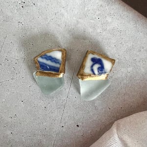 Può includere: Un paio di orecchini unici. Ogni orecchino presenta un pezzo di ceramica blu e bianca con bordi dorati, attaccato a un pezzo triangolare azzurro. Gli orecchini sono posizionati su uno sfondo grigio.