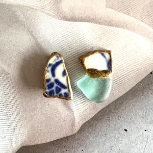 May include: A pair of earrings featuring irregular fragments of ceramic and glass. One earring displays a white and blue patterned ceramic shard, while the other combines a white and blue ceramic piece with a translucent light green sea glass.