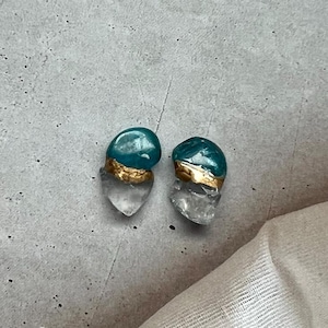 以下が含まれることがあります： 手作りのスタッドピアス。各イヤリングは、透明なハート型のクリスタルベース、ターコイズ色の丸い石、金色のバンドが特徴です。イヤリングは灰色の表面に表示されています。