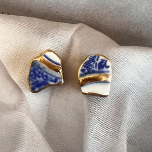 Könnte beinhalten: Ein Paar Ohrstecker aus unregelmäßigen Scherben von weiß-blau gemusterter Keramik. Jedes Stück weist ein glattes, goldfarbenes Metallic-Finish auf, das entlang der Kanten und über die Mitte der Keramikoberfläche aufgetragen wurde.