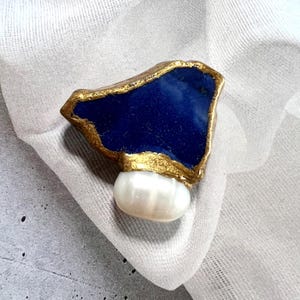 Könnte beinhalten: Eine Brosche mit einem großen, unregelmäßig geformten tiefblauen Stein mit einem strukturierten, goldfarbenen Metallrand. An der Basis des blauen Steins befindet sich eine einzelne, glatte, ovale weiße Perle mit einer glänzenden, irisierenden Oberfläche.