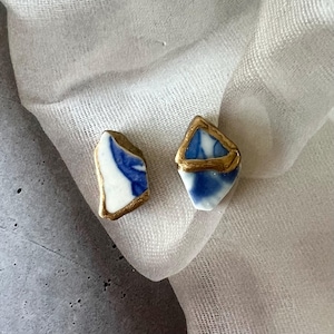 以下が含まれることがあります： 2つの手作りのイヤリング。各イヤリングはユニークな形で、白いベースに青い模様と金色の縁取りが施されています。イヤリングは白い布の背景に表示され、繊細なデザインと職人技を際立たせています。