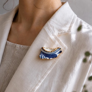 May include: A brooch crafted from a curved fragment of white ceramic featuring a blue painted pattern. The edges of the ceramic piece are finished with a metallic gold trim, creating a distinct contrast against the white and blue surface.