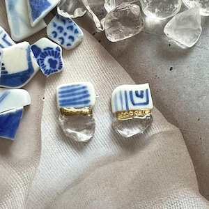 Puede incluir: Pendientes hechos a mano con piezas de porcelana blanca con rayas y diseños azules, acentuados con metal dorado y piedras transparentes de forma irregular. Los pendientes se muestran con otros fragmentos de porcelana y piedras transparentes.