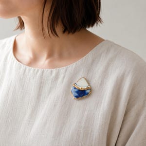 May include: A brooch featuring two irregular, layered ceramic shards with gold-toned edges. The top shard is solid white, while the bottom shard displays a blue and white marbled pattern. The piece is pinned to a light beige, textured linen fabric.