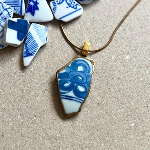 Puede incluir: Un collar con colgante que presenta un fragmento de cerámica blanca con patrones florales azules, engastado en un marco de metal dorado. El colgante cuelga de una fina cadena dorada, acompañado de varios fragmentos de cerámica azul y blanca sueltos.