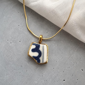 Puede incluir: Un collar de cadena de serpiente dorada con un colgante hecho de un fragmento de cerámica blanca con un patrón pintado en azul. La pieza de cerámica irregular está encerrada en un borde de metal dorado y cuelga de una sencilla anilla dorada.