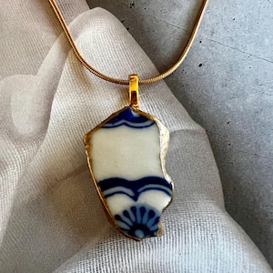 May include: A pendant necklace featuring a shard of white ceramic with blue floral and wave patterns. The ceramic piece is edged in gold and suspended from a thin gold-toned snake chain. The design highlights a smooth, irregular shape.