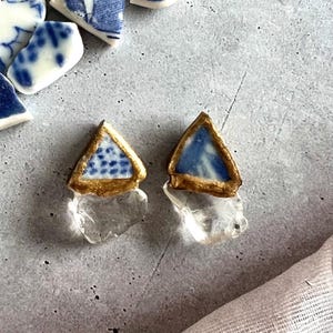 以下が含まれることがあります： 金色の縁取りが施された、三角形の青と白の磁器のトップと、透明なクリスタルのボトムが特徴の手作りのイヤリング。 イヤリングは灰色の背景に配置され、磁器の破片が散らばっています。
