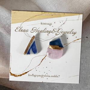 Könnte beinhalten: Ohrringe im Kintsugi-Stil. Ein Ohrring ist ein blau-weißes Dreieck mit goldenen Akzenten. Der andere ist eine rosa-blaue Form mit Gold. Die Ohrringe befinden sich auf einer weißen Karte mit goldenen Akzenten und dem Text "Elena Healing & Jewelry."