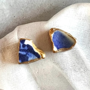 Puede incluir: Dos pendientes de botón con piezas planas e irregulares de cerámica azul y blanca. Cada pieza está enmarcada con un borde metálico de color dorado texturizado, resaltando la forma orgánica y dentada de los fragmentos.