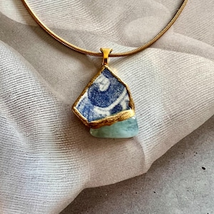 May include: A necklace featuring a gold-toned snake chain and a pendant composed of a blue and white patterned ceramic shard layered over a smooth, pale green stone, with both elements encased in a gold-colored metal border.