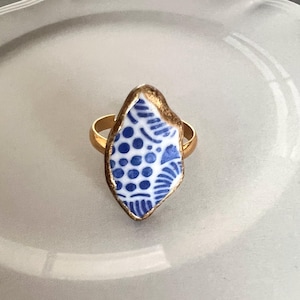 May include: A ring featuring a central shard of white ceramic with a blue pattern of dots and lines. The ceramic piece is framed by a metallic gold-toned border and is attached to a smooth, polished gold-toned metal band that encircles the finger.