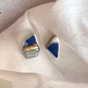 以下が含まれることがあります： 手作りのセラミックイヤリング2つ。 片方のイヤリングは、青とグレーの不規則な形で、金色のアクセントが付いています。 もう一方のイヤリングは、青と白の三角形で、金色のディテールが付いています。 イヤリングは白い布の上にあります。