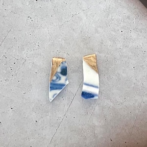Puede incluir: Dos pendientes hechos a mano con fragmentos de porcelana. Cada pendiente presenta una forma única con patrones azules y blancos y detalles de pan de oro. Los pendientes se exhiben sobre una superficie gris.