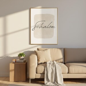 Op de afbeelding: Een ingelijste kunstdruk met het woord "Shalom" in elegant schrift. Het kunstwerk heeft een beige achtergrond in aquarel-stijl. De gouden lijst past bij de neutrale tinten van de print, die boven een beige bank met kussens en een deken hangt. Een houten bijzettafel met een plant en een kaars staat naast de bank.